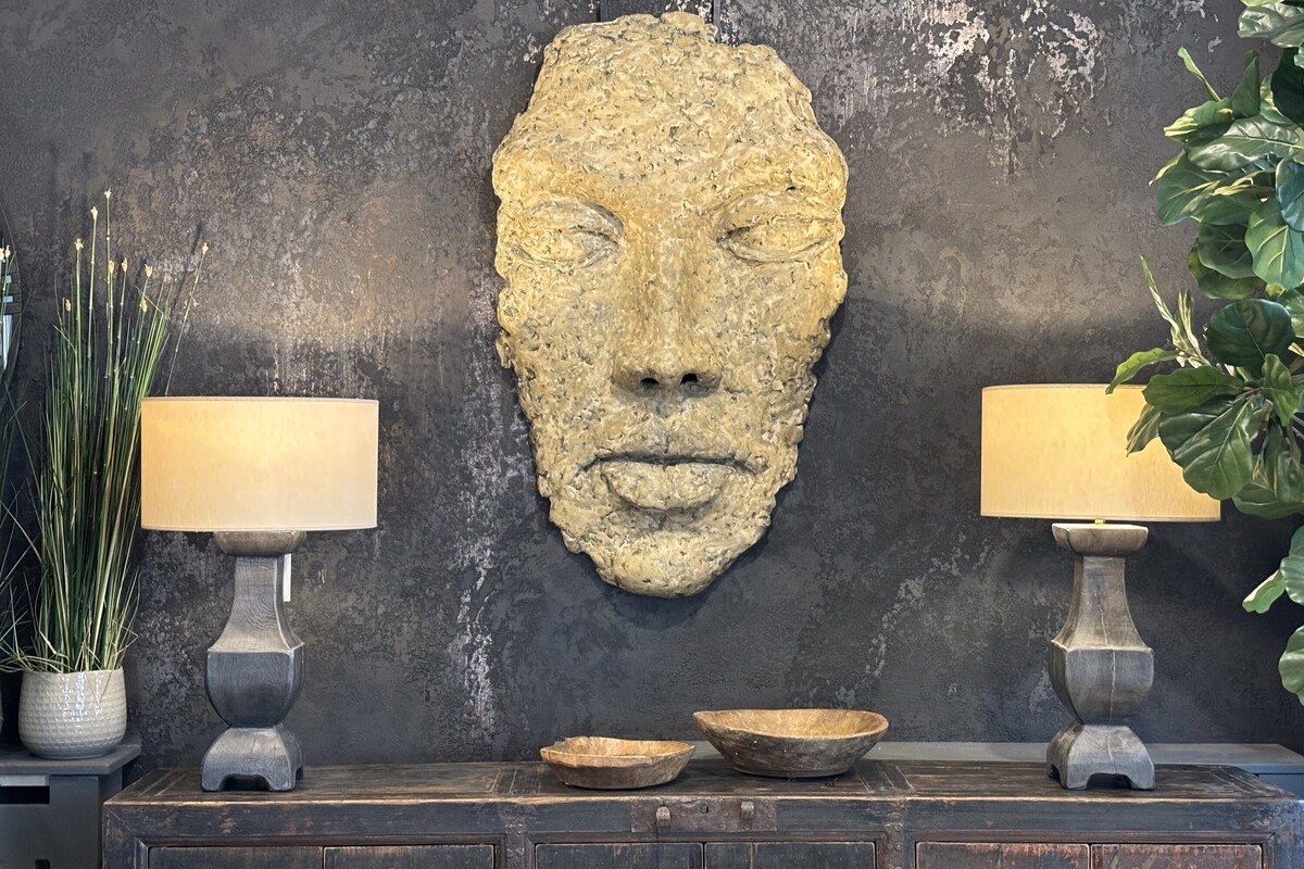Duża płaskorzeźba na ścianę w kształcie głowy. Maska w kolorze złoto-bezowym. Orygianlne drewniane lampy na tle czarnej ściany. Tynk dekoracyjny w kolorze czarno-złotym.