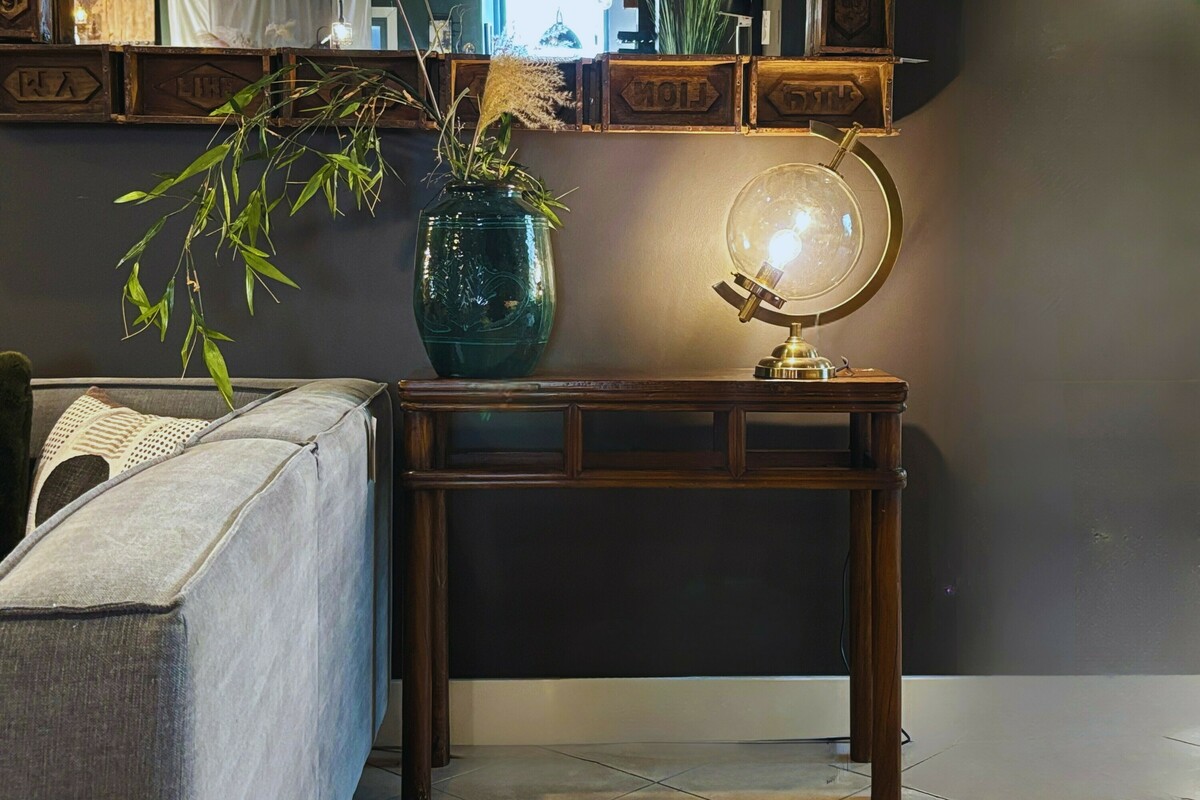 Mała drewniana konsola na czarnej ścianie. Konsolka do hollu lub wiatrołapu w małym rozmiarze. Zielony wazon ceramiczny, lustro w ciekawej ramie. Lampa globus,złota lampa w kształcie globusa.