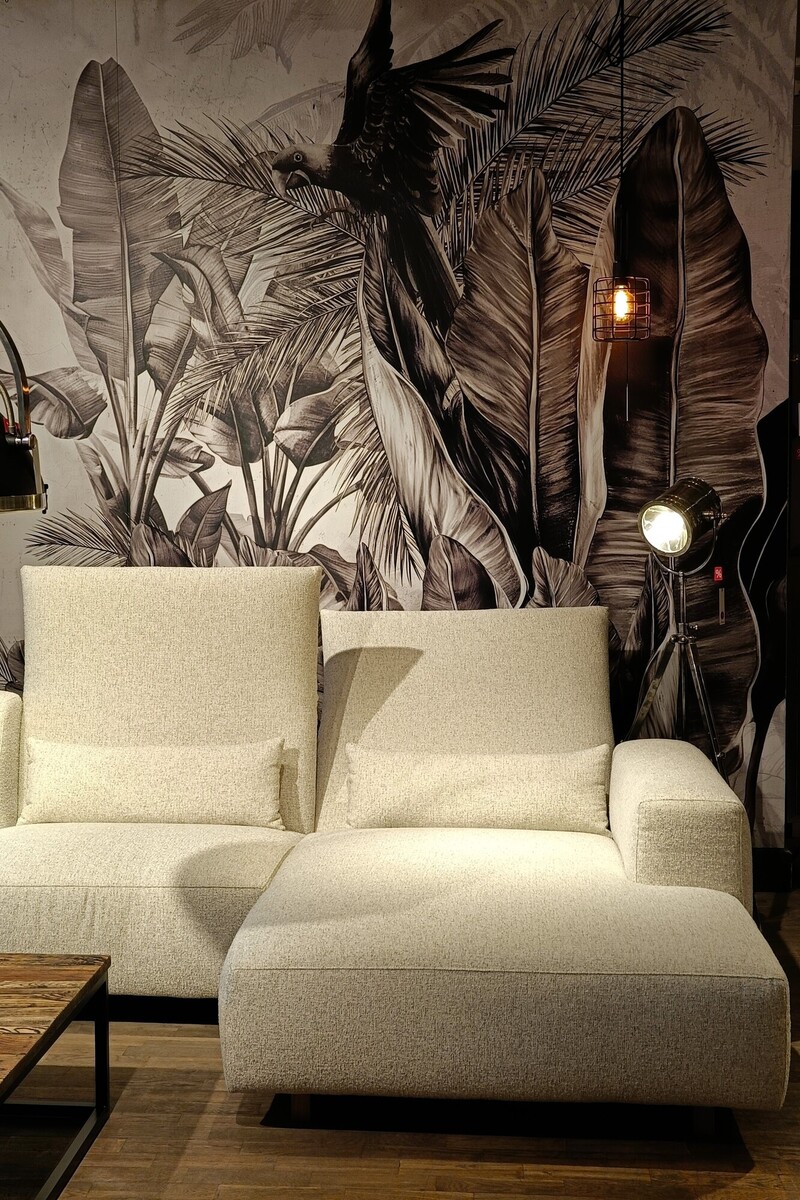 Szeroka kanapa z szezlongiem, lampa podłogowa reflektor, ściana w liście