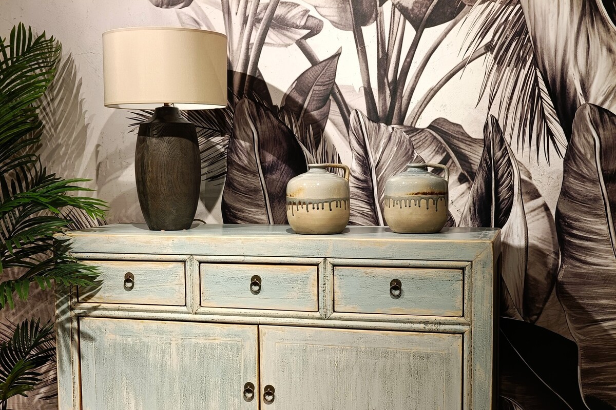 Błękitna komoda na tle tapety w liście, drewniana lampa z beżowym abażurem, szaro-niebieska ceramika dekoracyjna 