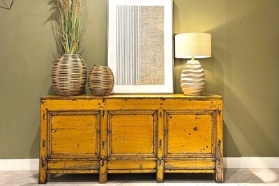 Duża żółta komoda na oliwkowej scianie. Piękna ryflowana, beżowa lampa z naturalnym abazurem. Trawa pampasowa w wazonie.